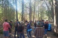 A large group of people outdoors in the woods hearing a lesson about wilderness navigation